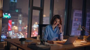 İş yerinde akşam saatlerinde cep telefonuyla danışmanlık. Gülümseyen iş kadını, akıllı telefon konuşmaları not defterine notlar yazıyor. Pozitif kadın yönetici, gece ofisinde telefonla sipariş veriyor..