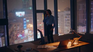 Siluet iş kadını panoramik pencerenin yanında cep telefonuyla konuşuyor. Şehir manzaralı. Zarif kadın yönetici gece geç saatlerde akıllı telefonla pazarlık yapıyor. Güvenilir işyeri sahibi telefonla görevi tartışıyor.