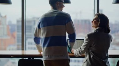 Meslektaşına katılan işçi, büyük bir pencerenin açık alanının arka görünümünde bilgisayar açıyor. Dost canlısı iki kadın iş arkadaşı laptoplarda çalışıyor ve kapalı mekanda iletişim kuruyor. Yaratıcı ajans uzmanları