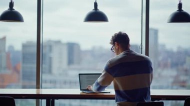 Bilgisayarı kullanan yaratıcı adam panoramik pencerenin önünde kahve içiyor. Şık bir yönetici şehir manzarası için ara verdi. Ciddi yaratıcı, modern gökdelendeki ofiste dinlenen fikirleri düşünüyor.