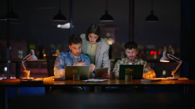 Kadın lider panoramik ofisteki çalışanlara danışmanlık yapıyor. Gece ekibi ortak çalışma alanında dizüstü bilgisayar kullanıyor. Akşamları çalışan bilgisayarları gözlemleyen bir yönetici. Proje süresi yaklaşıyor.