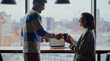 İş arkadaşı panoramik pencerelerdeki iş kadınına çay getiriyor. Şehir ofisindeki gayri resmi ortamda konuşan iş çifti. İşteyken flört eden iki yönetici şehrin arka planında mola veriyor