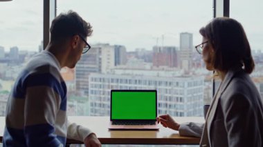 İki takım arkadaşı panoramik salonda çalışan yeşil ekran dizüstü bilgisayar görüntüsü veriyor. Yaratıcı ajans çalışanları bilgisayarın maketine bakıp kapalı mekanda iş kıyafetleri izliyorlar. Açık alanda oturan erkek kadın.