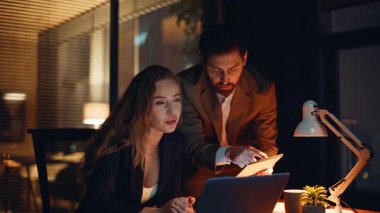 Birlikte iş yaptığı için mutlu iş arkadaşları. Gülümseyen kadın, akşam ofisinde bilgisayarla başarılı çalışma konusunu tartışıyor. Tableti kontrol eden iki iş arkadaşı kabinede ortaklığın keyfini çıkarıyor 