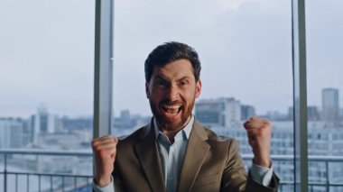 İş sahibi, kasvetli şehir manzaralı pencereye yakın mesafeden başarıya seviniyor. Heyecanlı duygusal yönetmen harika haberlere tepki veriyor. Kameraya odaklan. Neşeli acemi adam el kol hareketi yaparak para kazanıyor. 