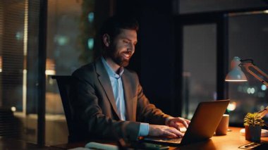 Çalışan meşgul bir adam, gece geç saatlerde iş yerinde defter çalışması yazıyor. Koyu ofiste laptopunu yudumlayan pozitif iş adamı. Gülümseyen patron çay içerek iş fikirlerini not ediyor.