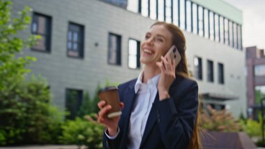 Neşeli girişimci cep telefonuyla sohbet ediyor kahve içiyor ve yakın plan çekiyor. Neşeli iş kadını cep telefonu sohbetinden hoşlanıyor modern ofislerin önünden geçiyor. İş kıyafetli gülümseyen kadın çağırıyor..
