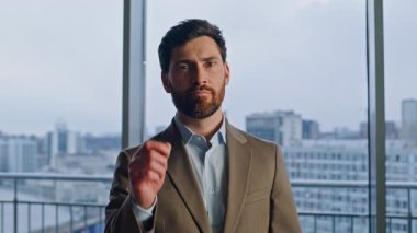 Gülümseyen CEO panoramik pencere ofis portresine parmağıyla işaret ediyor. Şirket binasında şık takım elbiseli mutlu bir işadamı duruyor. Başarılı bir işletme sahibi liderliğini ifade ediyor.