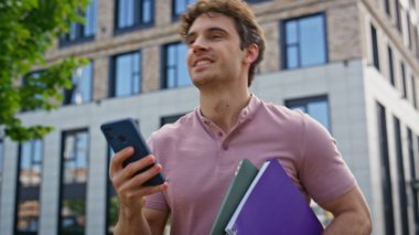 Modern binanın orada elinde akıllı telefon ve defterler tutan mutlu bir adam var. Kendine güveni olan bir adam, gülümseyen bir cep telefonu okuyarak şehir ortamında güneşli bir günde dolaşır. Neşeli öğrenci sohbeti.