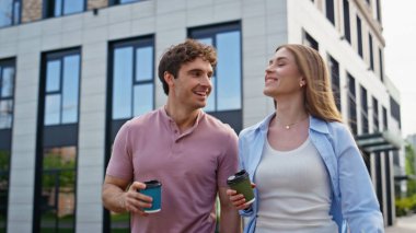 Rahatlamış iş arkadaşları dışarıda kahve siparişleriyle sohbet ediyorlar çağdaş binanın yakınında yürüyorlar. Genç arkadaşlar şehir merkezinde gezinmenin keyfini çıkarıyorlar. Gülümseyen kadın içeceklerden bahsediyor..