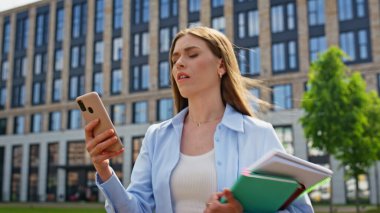 Meşgul iş kadını videosu, akıllı telefonu kapalı tutuyor. Elinde defterlerle gezen gülümseyen bir kadın şehir mimarisinin yanında yürüyüp telefon kamerasıyla konuşuyor. Kendine güvenen iş kadınının online toplantısı var.