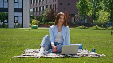 Gülümseyen kız videosu, güneş ışığında dizüstü bilgisayarlı yeşil çimen çimlerini arıyor. Neşeli kadın Green Park 'ta dostça sohbet ediyor. Bilgisayarın sanal konferansında konuşan esmer kadın..