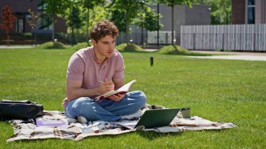 Çevrimiçi öğrenci videosu yeşil çim parkında oturan dizüstü bilgisayarı arıyor. Gevşemiş adam kitap yazarak Webinar 'ı güneşli doğada seyrediyor. Gülümseyen serbest çalışanın çimenli çayırda sanal toplantısı var..