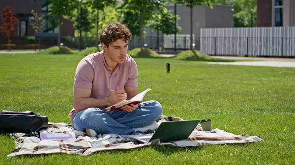 Çevrimiçi öğrenci videosu yeşil çim parkında oturan dizüstü bilgisayarı arıyor. Gevşemiş adam kitap yazarak Webinar 'ı güneşli doğada seyrediyor. Gülümseyen serbest çalışanın çimenli çayırda sanal toplantısı var..