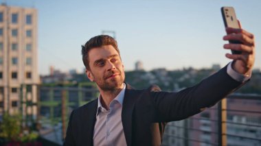 Günbatımı terasında şehir manzaralı cep telefonuyla selfie çeken şık bir işadamı. Gülümseyen iş adamı akıllı telefondan portre fotoğrafı çekiyor. Şehir manzarasının önünde telefon kamerası kullanan mutlu yönetici