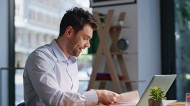 Modern ortak çalışma alanındaki dizüstü bilgisayarını kullanarak mutlu serbest çalışan videoları. Gülümseyen adam arkadaş canlısı online konferansta konuşuyor. Neşeli yöneticinin elinde kağıtlar var. Sanal sohbette anlaşma tartışıyorlar..