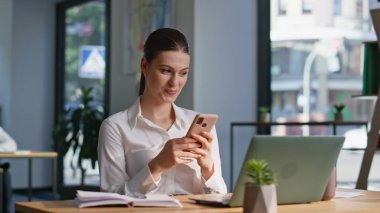 Ofisteki profesyonel mesajlaşma cep telefonu modern çalışma alanına yakın duruyor. Gülümseyen iş kadını, şirket çalışma masasında akıllı telefondan mesaj atıyor. İş yerinde cep telefonuyla sosyal medyayı karıştıran mutlu kadın.