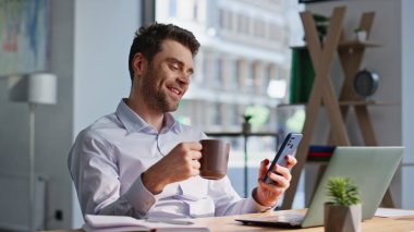 Ofisteki adam elinde kahve fincanı ile akıllı telefondan sosyal medyayı tarıyor. Gülümseyen girişimci iş yerinde cep telefonu mesajlaşması oturma masasını kırıyor. Mutlu yönetici, iş yerinde içki molası vermekten hoşlanır.