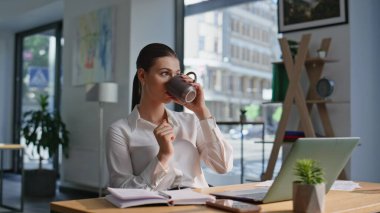 Rahat iş kadını kahve içiyor, modern iş yerinde mola veriyor. Ofiste dinlenen, sıcak içeceğini yudumlayan zarif bir kadın. Çekici bir bayan yönetici elinde fincan oturma masası tutuyor..