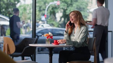 Samimi bir restoranda cep telefonuyla konuşan gülümseyen bir iş kadını. Kafeteryada cep telefonuyla konuşan mutlu güzel bir kadın. Neşeli bayan elinde kahve fincanıyla akıllı telefonu arıyor..