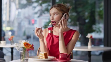 Lüks restoranda pasta yiyen kırmızı elbiseli bir kadını aramak. Cep telefonundan konuşan zarif bir bayan kafeteryada şeker tadıyor. Güzel kız akıllı telefondan konuşuyor, rahat bir kafe masasında oturuyor..