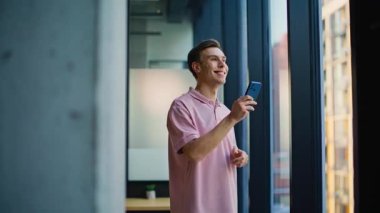 Hafif modern ofis kapanışında elinde cep telefonu alışverişi tutan memnun adam. Gülümseyen adam akıllı telefonuyla internette geziniyor. Mutlu girişimci mesaj cep telefonunu okuyor