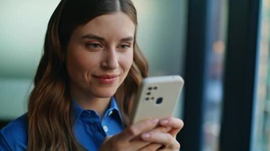 Kadının elleri cep telefonunu yazarken pencerenin önünde dikiliyor. Rahatlamış gülümseyen bayan internet mobil uygulamasından sipariş veriyor. Güzel iş kadını kapalı alanda akıllı telefondan online alışveriş yapıyor.