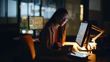 Geç dönem ekonomist, gece geç saatlerde iş merkezindeki masaüstü ekranında grafikleri analiz ediyor. Gözlüklü kadın tek başına oturmuş mali eğilimleri inceliyor. Adanmış uzmanlık alanı yatırım araştırması