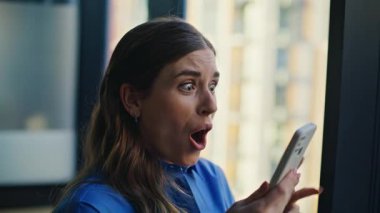 Cep telefonundan mesaj alan heyecanlı kadın duygusal olarak yakın çekim yapıyor. Akıllı telefondan harika haberler okuyan mutlu serbest çalışan kadın başarılı oldu. Pencerenin kenarında bekleyen çok mutlu bir bayan.