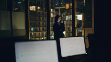 Panoramik iş birliğinde cep telefonu ekommerce uygulamasına göz atan gece işçisi. Neşeli kadın boş çalışma alanında dinlenen arkadaşından mesaj bildirimini kontrol ediyor. Akıllı telefondaki kendine güvenen sörf interneti