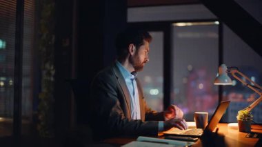 Gece apartmanında dizüstü bilgisayarda çalışan odaklanmış bir adam. Ciddi bir adam akşam evinde bilgisayar izlerken notları kontrol ediyor. Çevrimiçi yönetici e-postaya uzaktan cevap veriyor. Uzaklık iş konsepti 