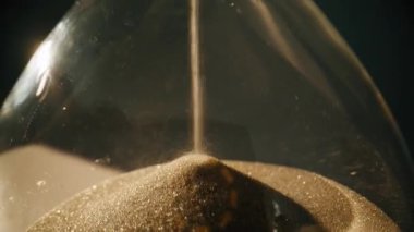 Macro view elegant hourglass in evening light. Closeup gold sand flowing inside transparent glass clock in super slow motion. Vintage timepiece measuring time indoors. Retro device for countdown.