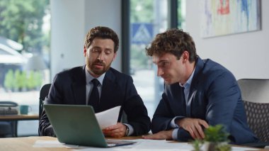 Gülümseyen meslektaşlar, modern ofis alanında dizüstü bilgisayar görüntülerini selamlıyorlar. Şirketin iş yerinde online takım buluşmasında konuşan iki pozitif adam. Üretken uzaktan beyin fırtınası sağlayan iş arkadaşları