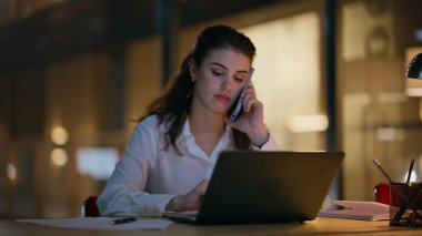 İş yerinde cep telefonuyla konuşan profesyonel bir kadın. Yakın plan. Ciddi bir iş kadını dizüstü bilgisayarla akşam ofisinde akıllı telefonu arıyor. Bayan akıllı telefon konuşmasında projeyi tartışır geç saatlere kadar çalışır
