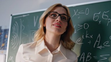 Üniversite öğretmeni, karatahtaya yakın çekimde fizik formüllerini gösteriyor. Gözlük takan gülümseyen kadın, bilinmeyen öğrencilerle etkileşime geçip cebiri açıklıyor. Dost canlısı eğitimci ders hakkında konuşuyor.