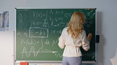 Matematik öğretmeni tahtaya denklemler yazıyor. Yakın plan. Lisede matematik öğretmenliği yapan profesyonel bir kadın eğitimci bilinmeyen öğrencilerle konuşuyor. Zarif bayan matematik formüllerini karatahtayla açıklıyor..