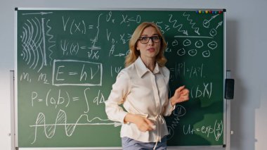 Bilinmeyen üniversite öğrencilerine matematik formüllerini açıklayan bir kadın. Gözlüklü gülümseyen öğretmen tahtaya matematiksel denklemler yazıyor. Okulda öğretmenlik yapan pozitif kadın eğitmen.