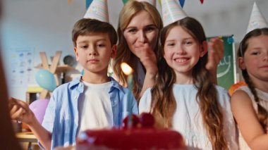 Çocuklar doğum günü partisini öğretmen ve renkli dekorasyonlarla kutluyorlar. Gülümseyen kız pasta mumları üflüyor, heyecanlanıyor. Mutlu çocuklar şapka takıp eğleniyorlar..