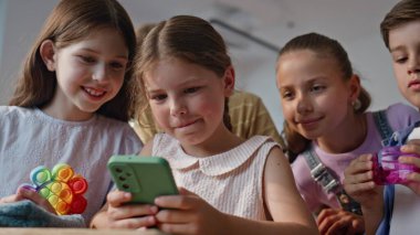 Sınıf arkadaşları sınıf odasında cep telefonlarına bakıyor. Okul arkadaşlarıyla akıllı telefon oyunu oynayan küçük kız sınıfta tezahürat yapıyor. Gülümseyen ilkokul öğrencileri telefonla eğlenceli etkileşimlerden hoşlanırlar