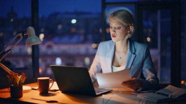 Çalışan meşgul bir kadın, gece geç saatlerde iş yerinde doküman okuyor. Karanlık ofiste dizüstü bilgisayar yazan ciddi bir iş kadını. Akıllı serbest çalışan bilgisayardaki iş fikirlerini not alıyor 