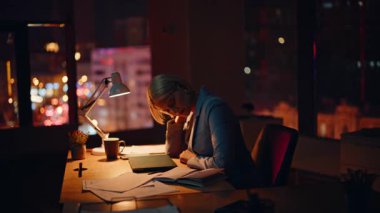 Yorgun bir ofis çalışanı, karanlık çalışma alanındaki çalışma masasında bitkin bir şekilde oturuyor. Hüzünlü iş kadını fazla mesai yapmak için nedensiz yere boynuna masaj yapıyor. Mutsuz sarışın gece gündüz çalışmaktan yoruldu.