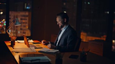 Geç dönem yöneticileri laptopları modern iş ofislerinde otururken görüntülüyor. Duygusal iş adamı, gece odasında çevrimiçi konferansta konuşuyor. Heyecanlı adam sanal buluşmada iş başarısını paylaşıyor