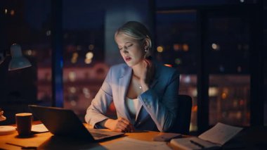 Yorgun bir ofis çalışanı, karanlık çalışma alanındaki çalışma masasında bitkin bir şekilde oturuyor. Üzgün iş kadını, akıllı telefon saatini kontrol ediyor. Fazla mesai yapmak istemeyen boynu ovalıyor. Mutsuz sarışın yorucu bir iş günü.