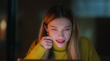Gece vakti dizüstü bilgisayarla sohbet eden bir kadın. Gülümseyen sarışın kadın, bilgisayar ekranı görünümlü, karanlık dairede oturuyor. Gece geç saatlere kadar ofiste çalışan çalışkan bir iş kadını. Gece işi kavramı.