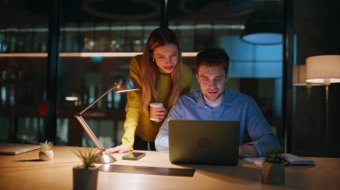 Karanlık ofis alanında dizüstü bilgisayara bakan gülümseyen ortaklar. Mutlu iki yönetici, gece kahve fincanları tutarak başarılı bir projeyi kutluyorlar. Neşeli meslektaşlar birlikte yaratıcı girişimi tartışıyor..
