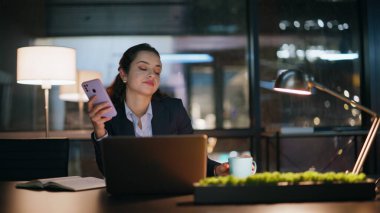 Ofisteki kadın gece akıllı telefon kullanıyor. Kapalı kapılar ardında kendini yorgun hissediyor. Rahat iş kadını cep telefonu mesajları okuyor. Koyu iş yerinde çay içiyor.