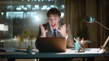 Heyecanlı iş adamı duygusal açıdan başarılı bir projeye tepki veriyor. Aşırı mutlu adam karanlık işyerinde dizüstü bilgisayar ekranına bakıyor.