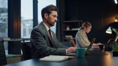 Akşam yöneticileri ofis iş yerinde dizüstü bilgisayarları klavyeyle kapatıyor. Modern işyerindeki bilgisayarlarda çalışan iki çalışan. Ciddi sakallı adam e-posta okuyor. Koyu dolapta kahve fincanı alıyor.