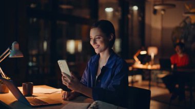 Freelancer cep telefonunda sohbet ediyor. Gece mesai saatleri kapalı. Gülümseyen kadın yönetici şirket işyerinde mesai yaparak mesai yapıyor. Cep telefonunu izleyen mutlu iş kadını