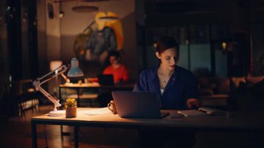 Karanlık ofiste gece vardiyasında çalışan iş arkadaşları. Profesyonel kadın yönetici dizüstü bilgisayarına göz atıp lamba ışığında notlar yazıyor. Başarılı bir proje üzerinde çalışan gülümseyen bir çalışan. İş ortaklığı